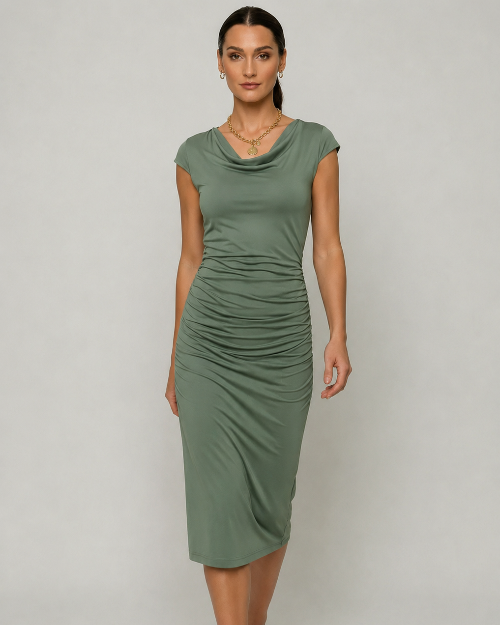 A woman modeling a sage green cowl neck midi dress with cap sleeves and ruched detailing at the waist, paired with black pointed-toe heels against a light gray studio background.