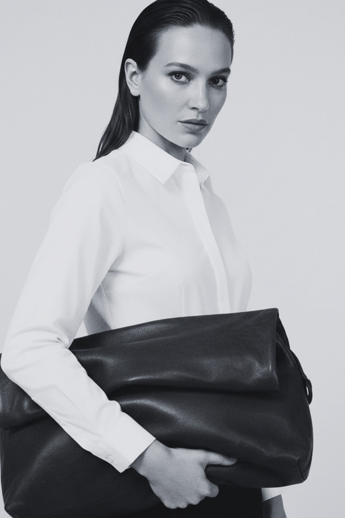 A woman in a white collared shirt and dark trousers holds a large black leather duffel bag against a light gray studio background.