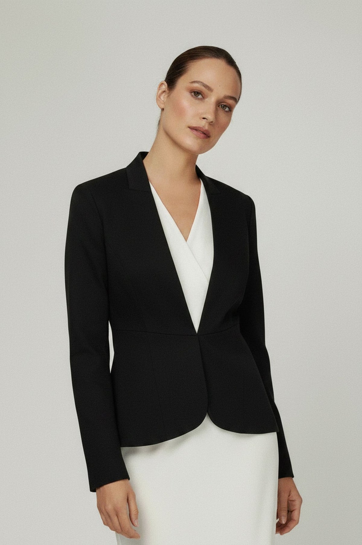 A woman modeling a black tailored blazer over a white V-neck top and white skirt, photographed against a plain light gray studio background.