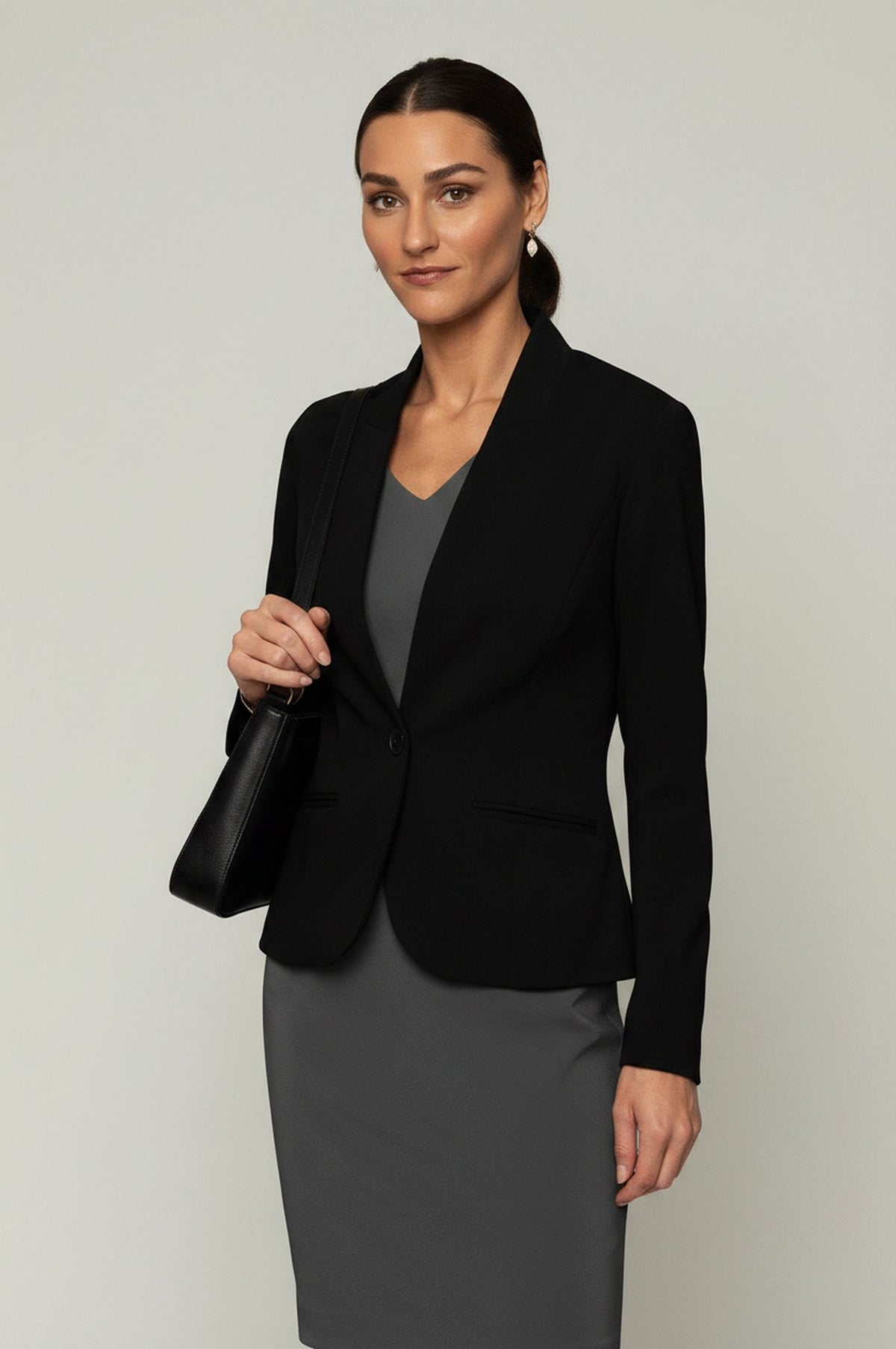 A woman modeling a black tailored blazer over a gray V-neck top and gray skirt, holding a black handbag against a plain light gray studio background.