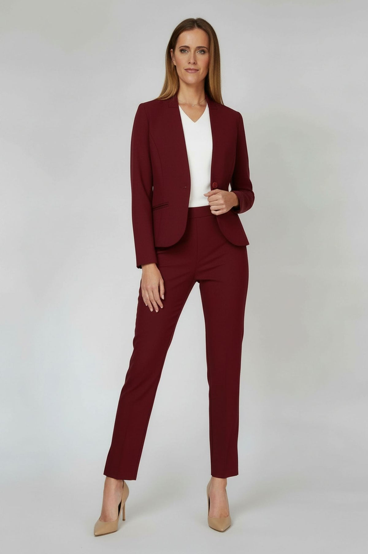 A woman modeling a burgundy two-piece blazer and trouser suit with a white top and beige heels, standing against a light gray studio background.