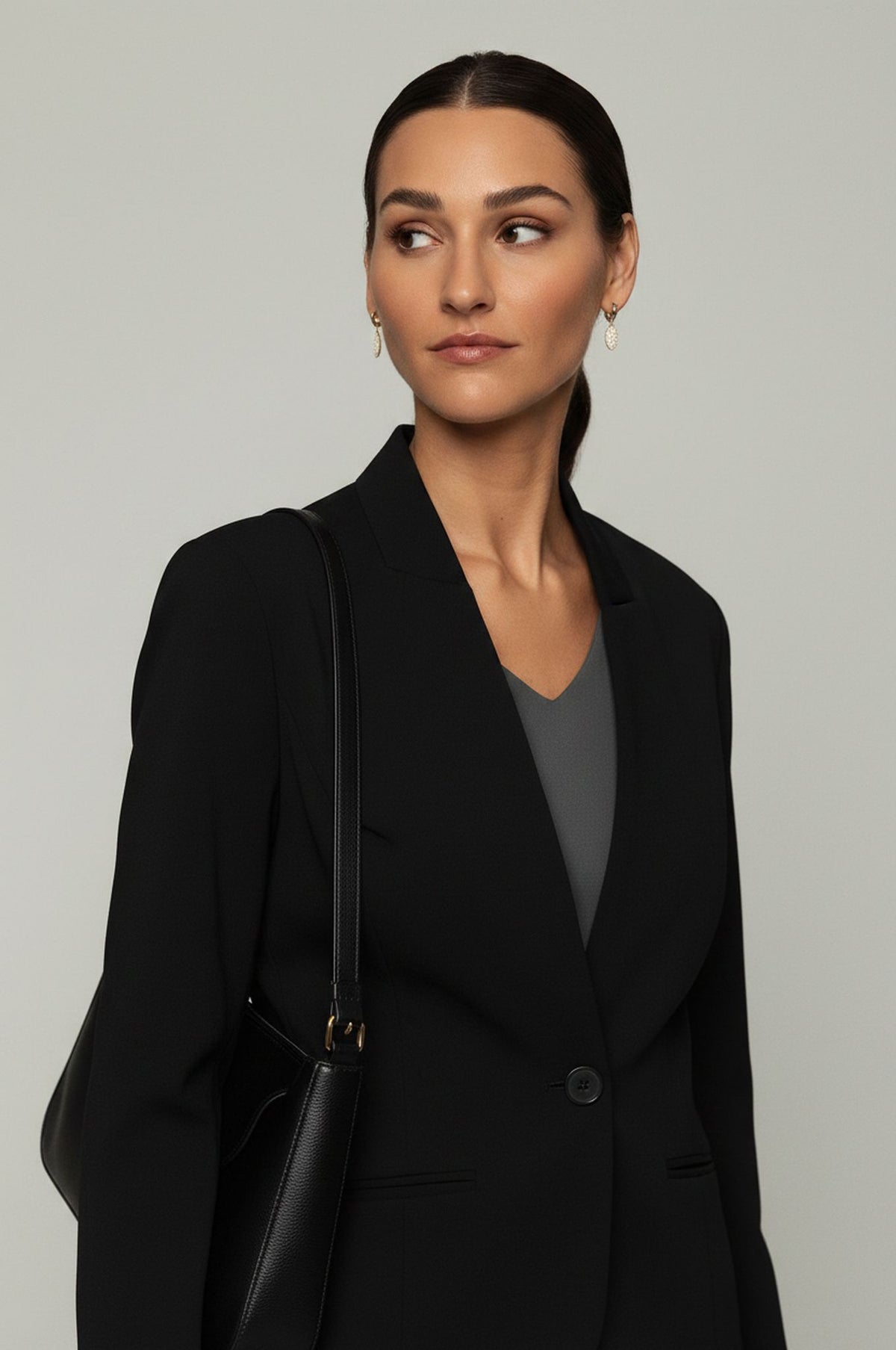 A woman wearing a black tailored blazer over a dark gray V-neck top, carrying a black shoulder bag against a plain light gray studio background.