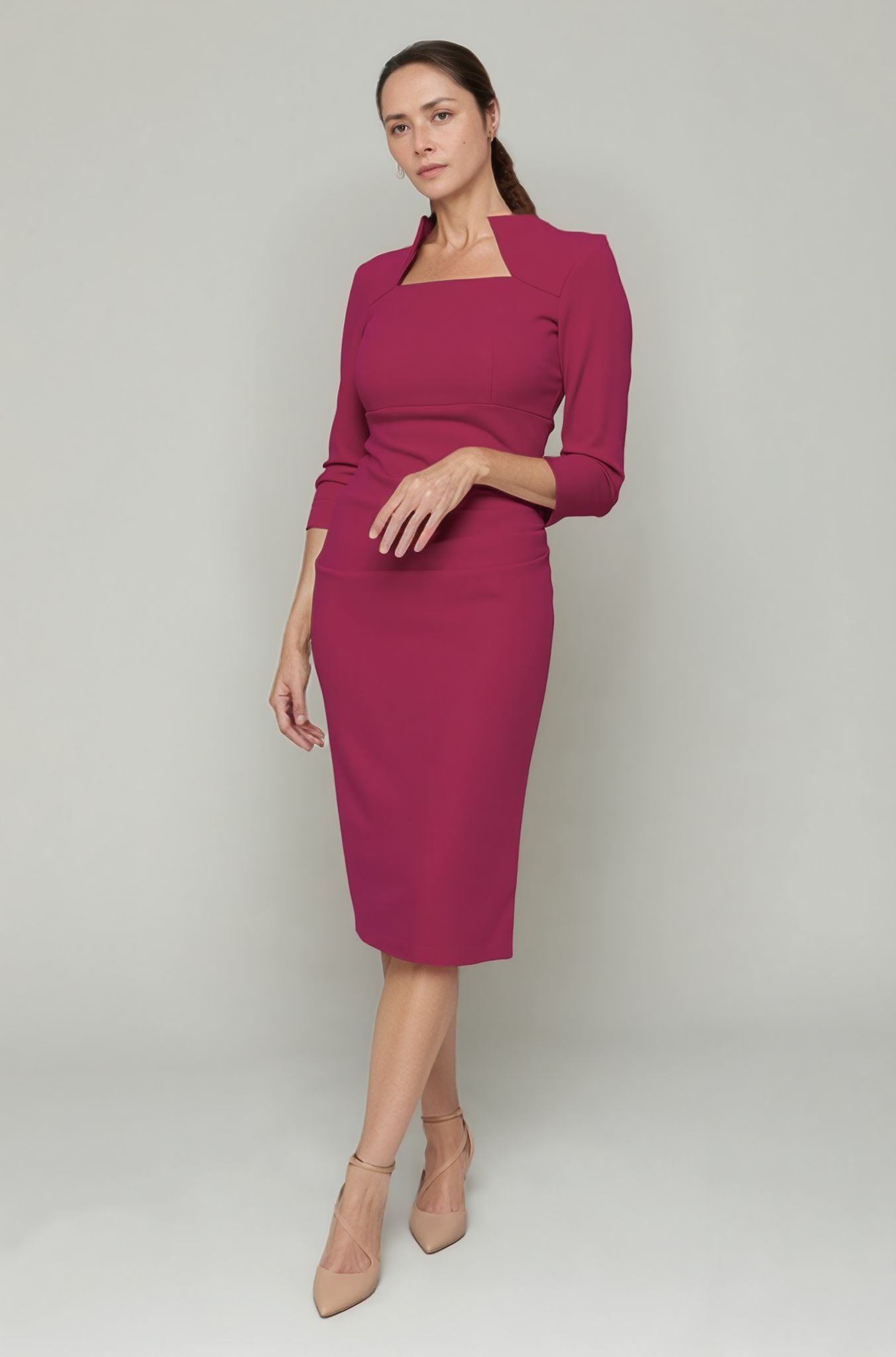 Woman with dark hair tied back wearing a pink magenta midi dress with a unique square neckline and subtle stand collar. The dress features three-quarter sleeves and elegant draping on the waist, creating a flattering silhouette. She completes the sophisticated look with nude strappy heels and minimalist earrings, exuding a polished and professional style.